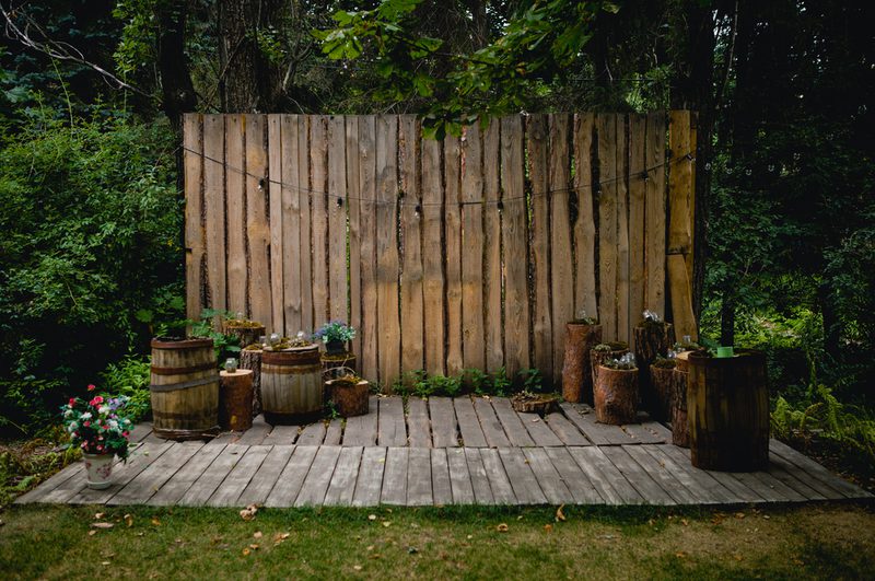 Rustic outdoor wooden stage with a vertical plank backdrop, string lights, barrels and stumps, and potted flowers on the left amid greenery.