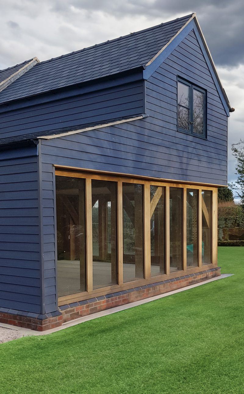 Blue two-story wooden house extension with large glass doors framed in natural wood, brick base, green lawn, cloudy sky.