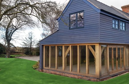 Blue two-story house with a large wooden-framed glass conservatory along the ground floor, brick foundation, and a bright green lawn with leafless trees.