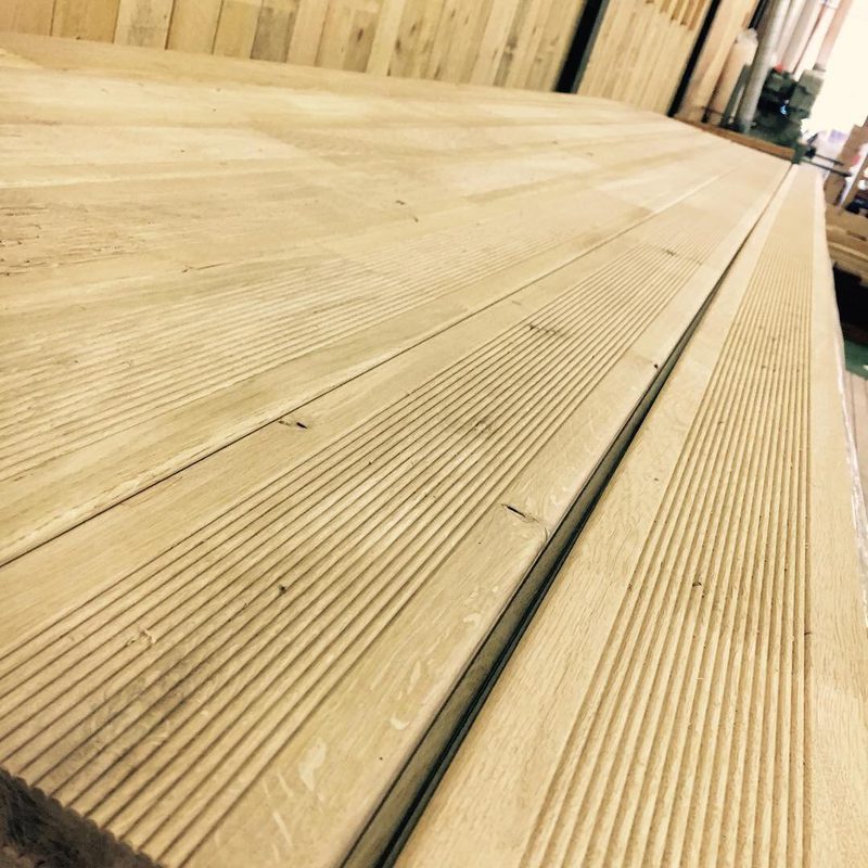 Close-up of light wood planks with prominent grain and grooves, laid in a long surface; a seam runs along the length, with a wooden wall in the background.
