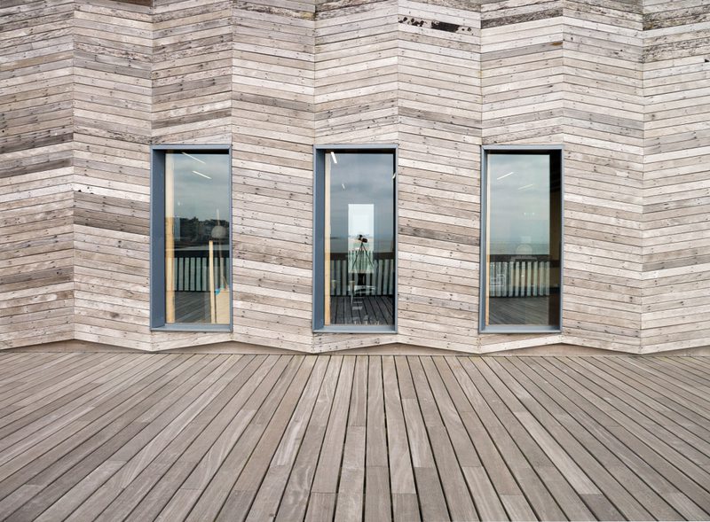 Three tall, narrow windows in a chevron wood-clad wall, gray-framed, with a wide wooden deck in front.