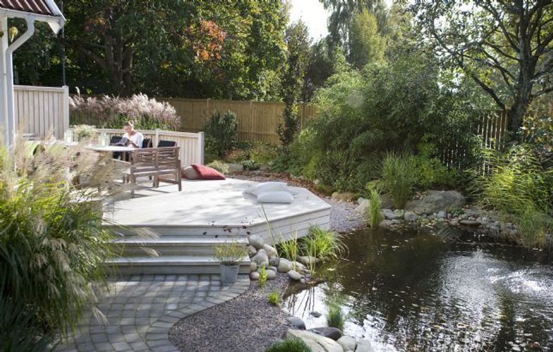 Backyard pond with a stone edge and wooden deck; two people sit at a table on the deck, surrounded by lush plants, trees, and a wooden fence.