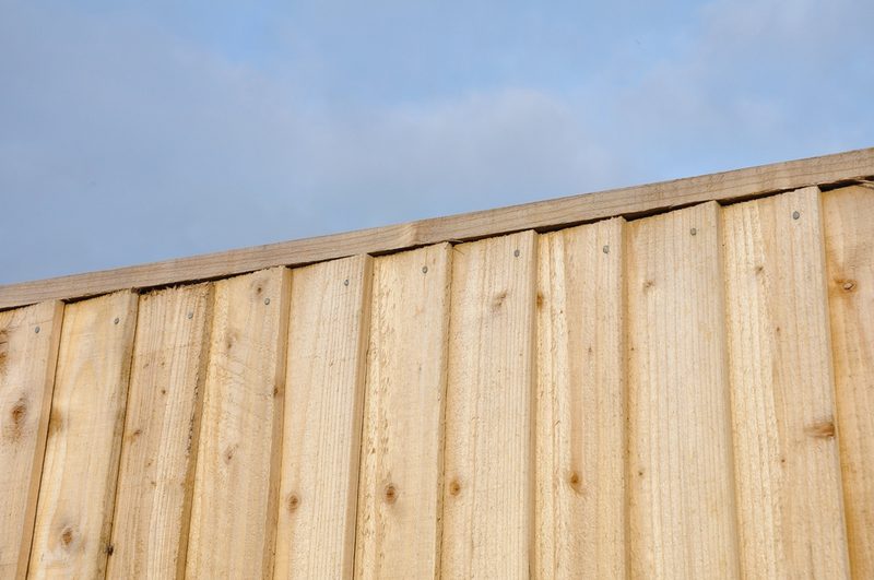 Vertical pale wood fence panels with visible grain and nails, topped by a horizontal rail, against a blue sky with a few clouds.