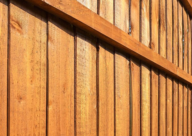 Close-up of a wooden fence with vertical planks in warm brown tones, a diagonal crossbeam across the panels, and visible wood grain and shading.