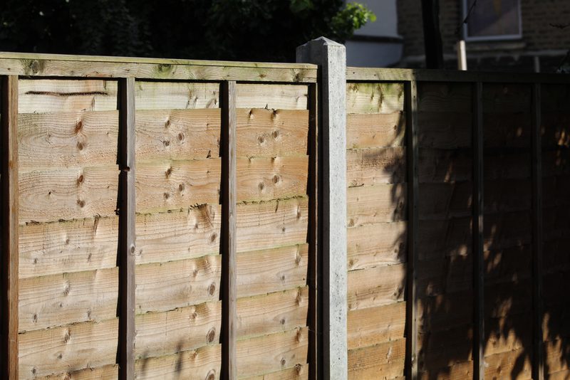 Wooden fence panels with vertical boards and a central concrete post, sunlit on the left and shadowed on the right; background shows trees.