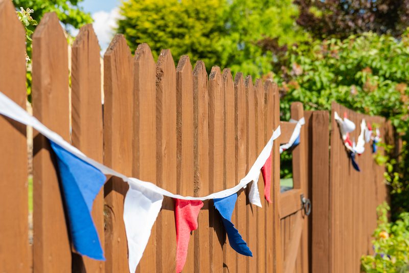 Brown wooden picket fence with blue, white, and red pennant bunting along a sunny garden backdrop.