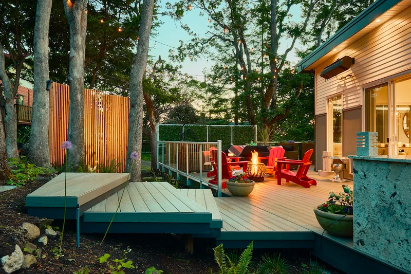 Evening backyard deck with string lights, a lit fire pit and red Adirondack chairs, potted plants, a fence, and a house with a counter on the right.