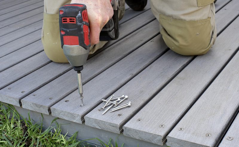 Close-up of a person kneeling on gray wooden deck boards, using a cordless drill to drive screws into the boards; a small pile of screws rests nearby.
