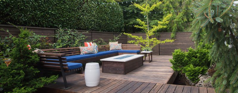 Outdoor wooden deck with an L-shaped blue-cushioned bench, a central fire pit table, patterned throw pillows, a white barrel, potted plants, and string lights along a wooden fence.