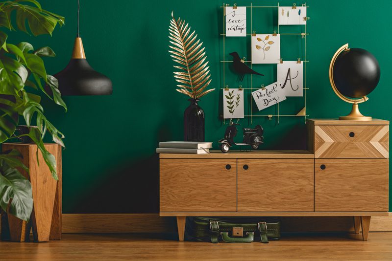 Mid-century wooden sideboard against a green wall with globe, art-card grid, pendant light, leafy plant, palm sculpture, books, bike, and suitcase underneath.