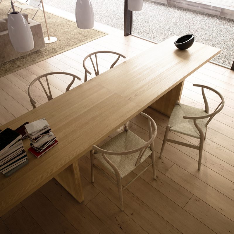 Long light wood table with white wicker chairs in a bright minimalist dining area; magazines stacked at one end and a small black bowl on the tabletop.