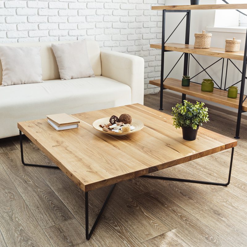 Bright living room with a white brick wall, beige sofa, and a large wooden coffee table on black metal legs; plant, books, and shelving with baskets.