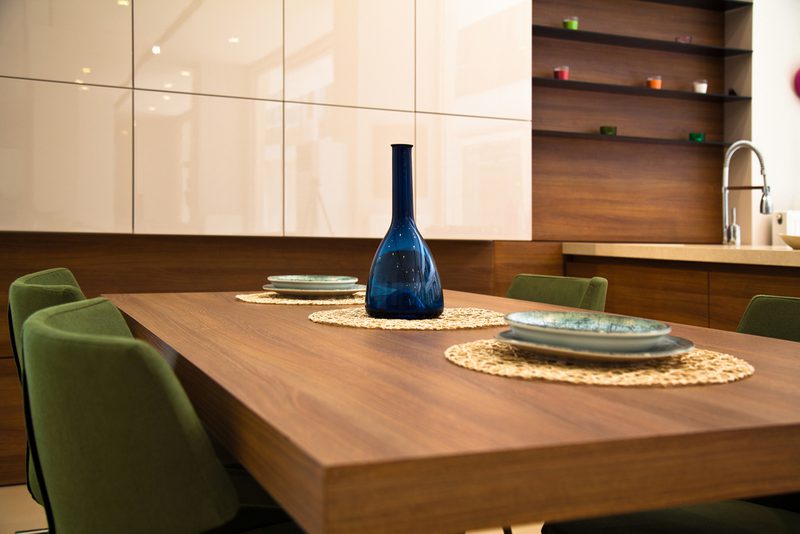 Wood dining table with a blue glass bottle centerpiece on woven placemats, green chairs, and white glossy cabinets in a modern kitchen.