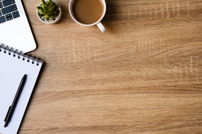 Wooden desk with a partial laptop, a spiral notebook with a black pen, a small succulent, and a white mug of coffee.