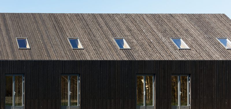 Modern dark wooden building with a long sloped roof and several skylights; ground level features tall glass doors and narrow windows under a clear blue sky.