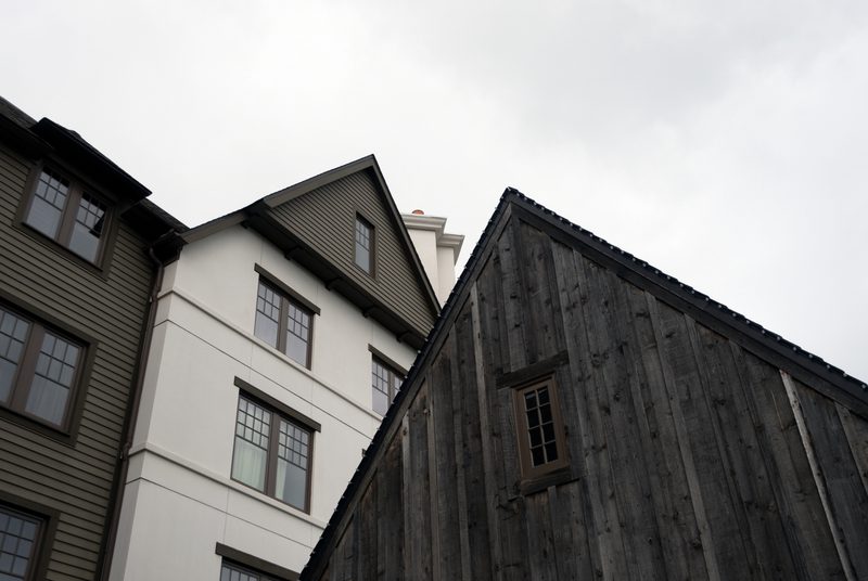 Three adjacent buildings from a low angle: brown clapboard left, white center building, and a weathered gray wooden shed on the right under a cloudy sky.