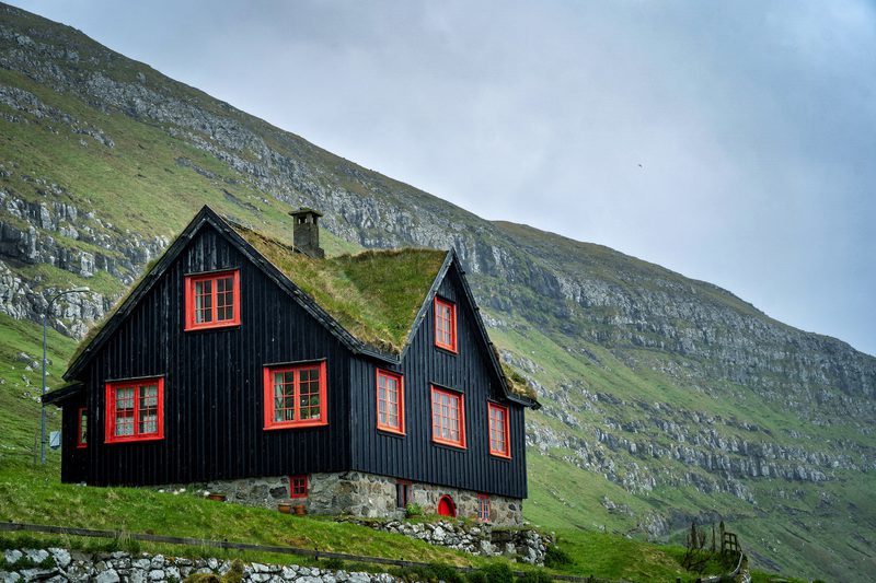 Dark blue wooden cottage with red-framed windows sits on a stone foundation, its grassy sod roof on a green hillside, with rocky mountains and a cloudy sky behind.
