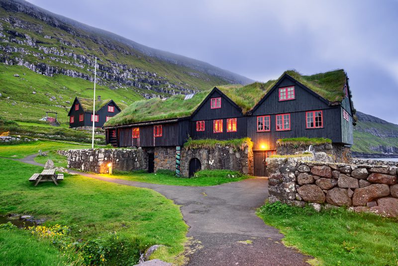 Dark wooden houses with sod roofs and red-framed windows along a stone wall, warm lights glow inside as green hills and rocky cliffs rise behind, with a path and a picnic table in the foreground.