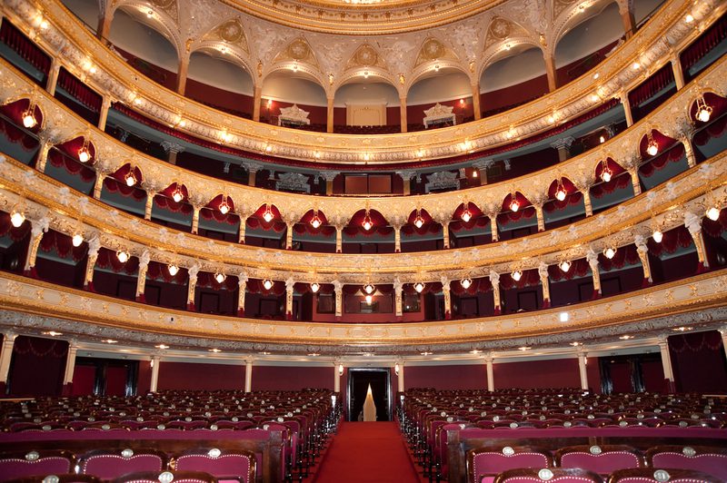 Opulent theater interior with gilded multi-tier balconies, red upholstered seats, and warm chandeliers, viewed from the center aisle.