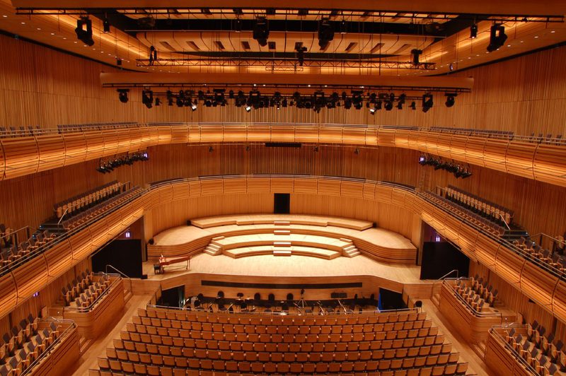 A large, wood-paneled concert hall with curved balconies surrounding a stepped, central stage, warmly lit by overhead lighting rigs.