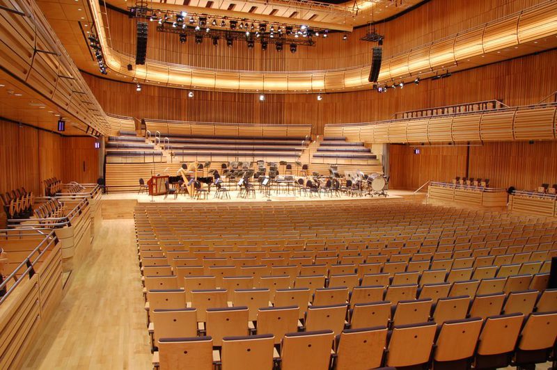 A wood-paneled concert hall with rows of empty seats facing a stage where an orchestra rehearses under warm overhead lights.