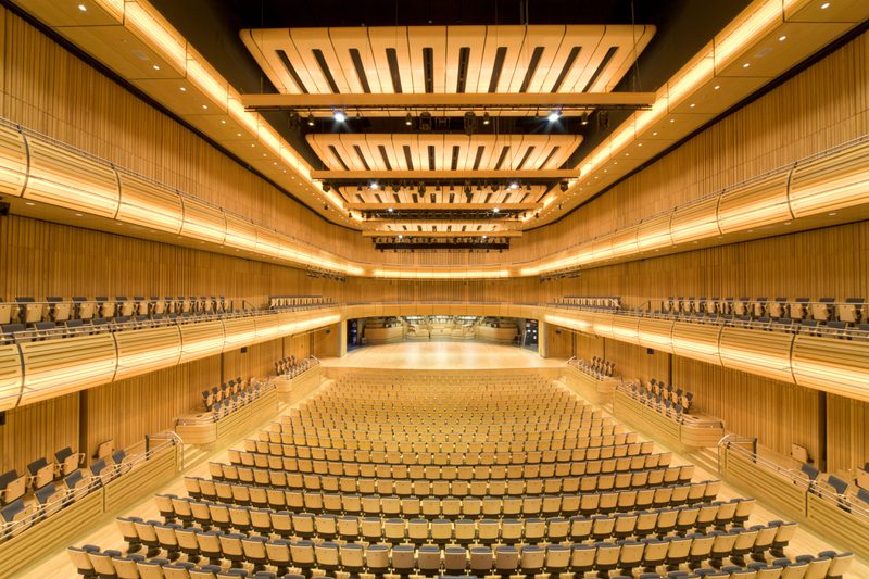 Empty wood-paneled concert hall with multiple seating tiers and a softly lit central stage; warm amber lighting and orderly rows of seats.