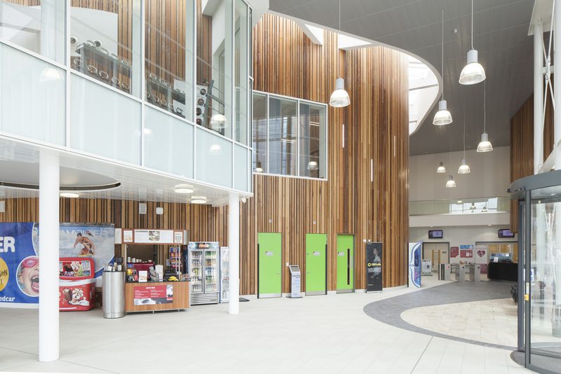 Modern atrium with wooden panels, glass balcony, columns, pendant lights, a snack bar with vending machines, lime green doors, and a curved floor.