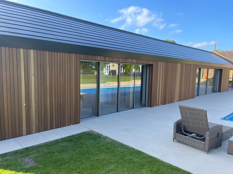 Single-story wood-clad building with vertical panels, large glass sliding doors, a gray tiled patio, wicker seating, and a swimming pool under a blue sky.