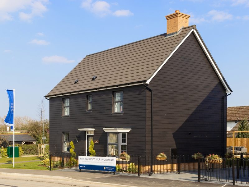 Two-story dark house with white trim, gray pitched roof, and a brick chimney; fenced front yard with planters and a sign under a blue sky.