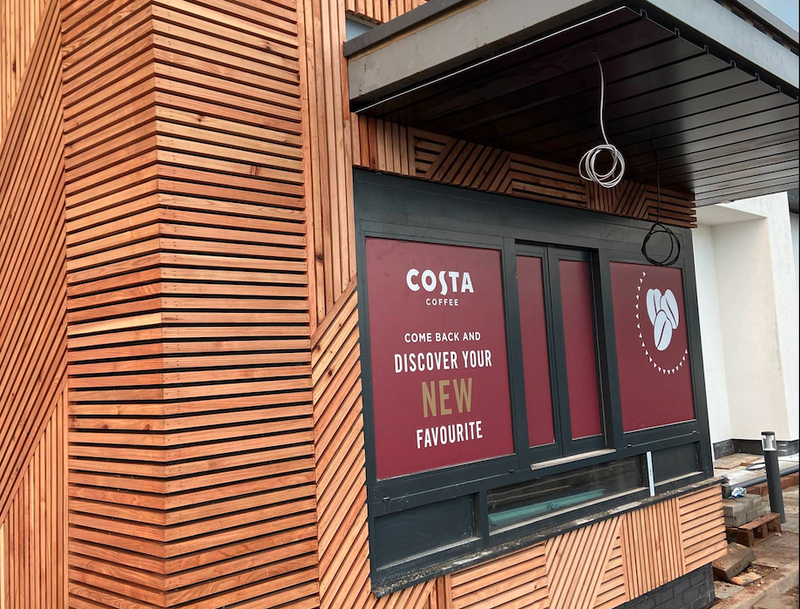 Exterior of a Costa Coffee store with a warm wooden slat facade, burgundy window panels, and a sign inviting you to discover your new favourite.