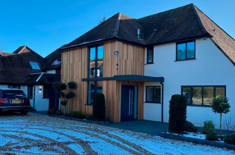 Modern two-story house with wooden cladding and white walls, blue front door, trimmed shrubs, and a car on a snowy gravel driveway under a blue sky.