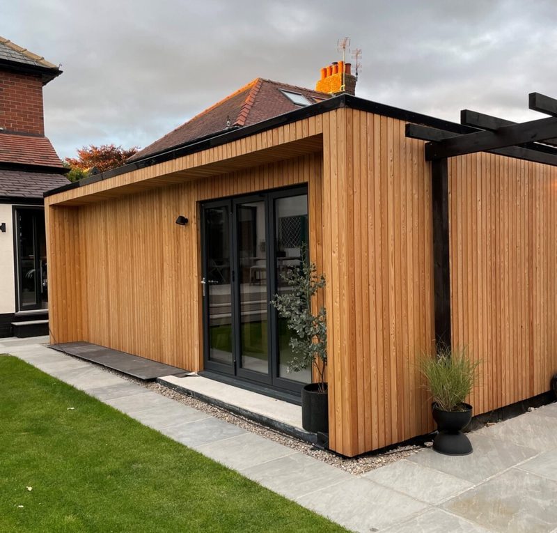 Wooden-clad single-story extension with large black-framed glass doors, a black pergola frame, potted plants, and a gravel strip by the green lawn.