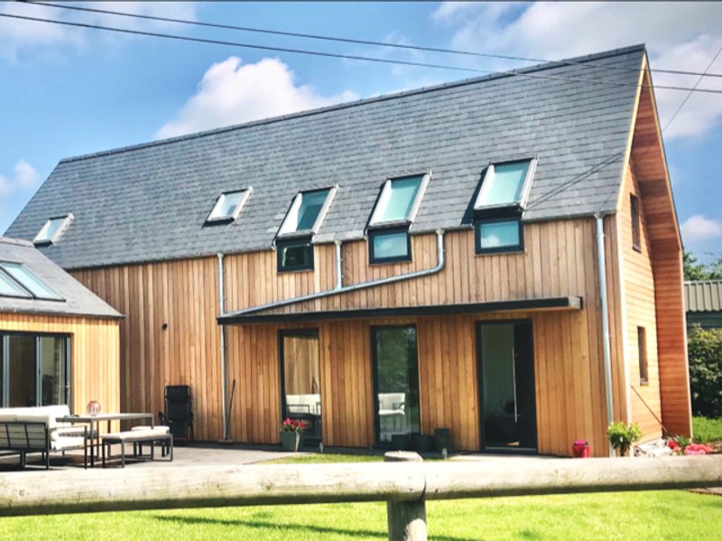 Modern wooden house with a dark slate roof, skylights, and glass doors opening onto a wooden deck with outdoor seating on a green lawn.