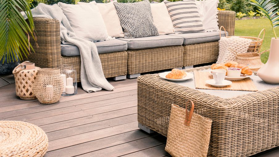 Outdoor wicker sofa set on a wooden deck with neutral cushions, a matching coffee table, baskets, and lanterns; croissants and tea on the table.