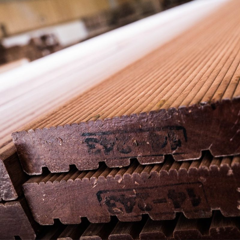 Close-up view of stacked brown wooden boards with grooved edges and visible grain, showing rough-cut ends and a blurred workshop-like background.