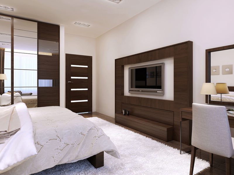 Modern hotel room with dark wood furniture, wall-mounted TV in a wood frame, a white rug, a desk with lamp, mirrored wardrobe, and a bed with a duvet.