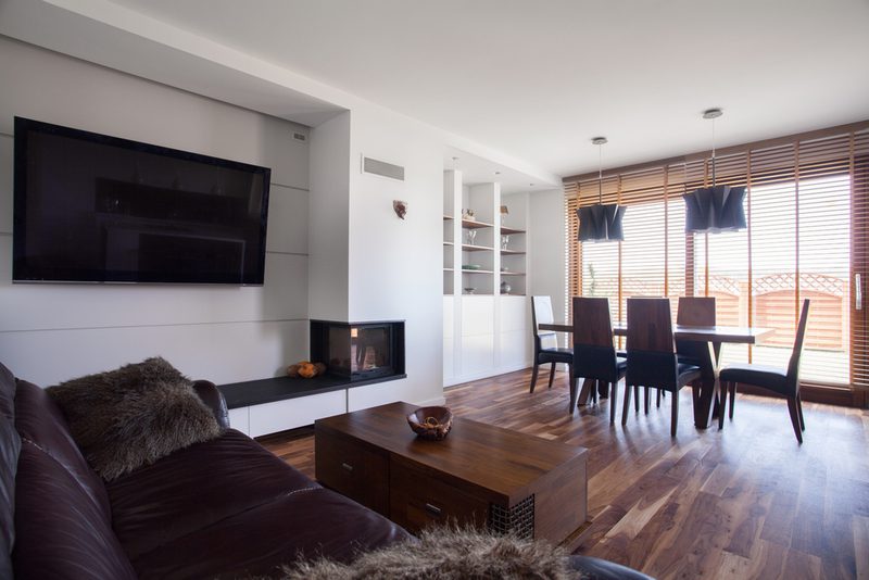 Modern living area with a wall-mounted TV above a fireplace, a dark brown sofa, wooden coffee table, white built-in shelves, and a dining set by glass doors with blinds.