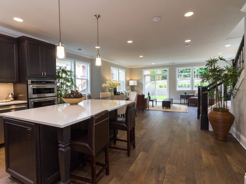 Open-concept kitchen and living area with dark wood cabinets, a white island, pendant lights, and a seating area by large windows and plants.