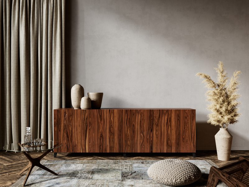 Minimal living room with a walnut sideboard, ceramic vases, tall beige curtains, glass coffee table, a woven pouf, pampas grass in a vase, and a neutral rug.