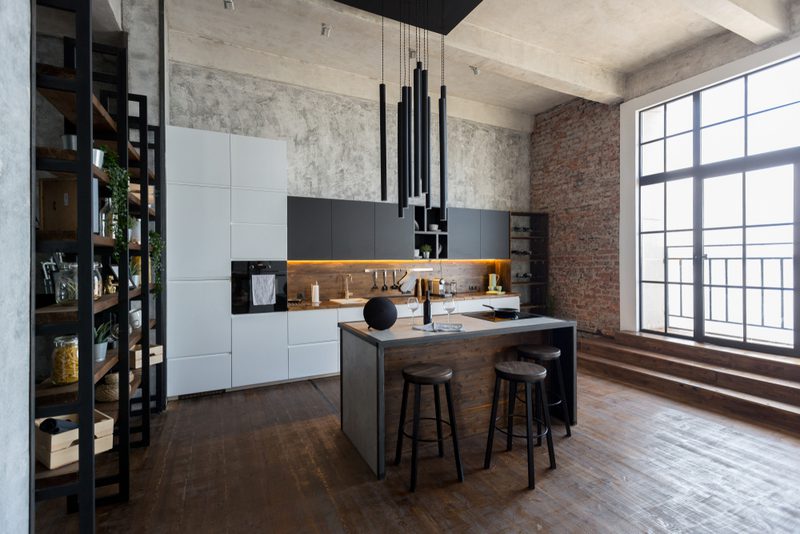 Industrial loft kitchen with exposed brick and a large window. Central island with a dark wood base and gray top, three black stools and pendant lights.