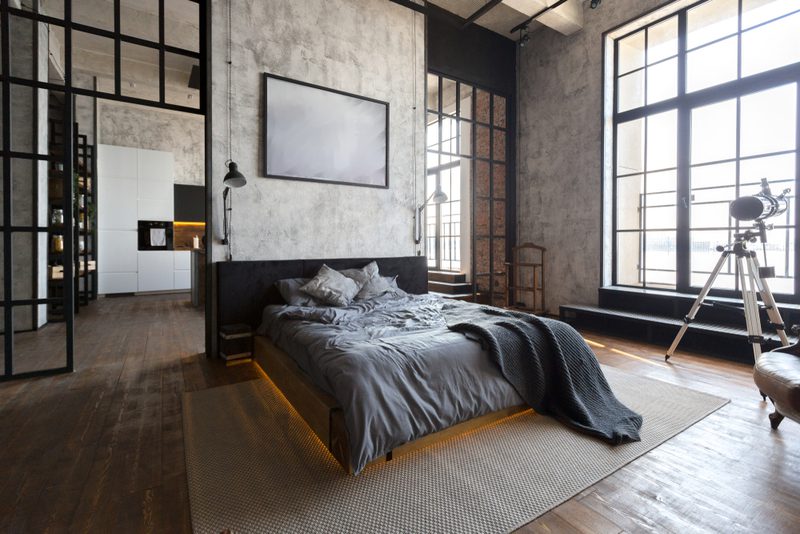 Industrial loft bedroom with exposed concrete walls, grid windows, and a low platform bed in gray linens. A tripod telescope sits by the window.