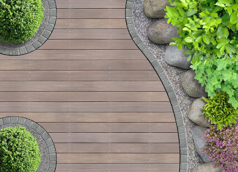Top-down view of a curved wooden deck bordered by gray stones, with lush green shrubs, rocks, and purple flowering plants along the right edge.