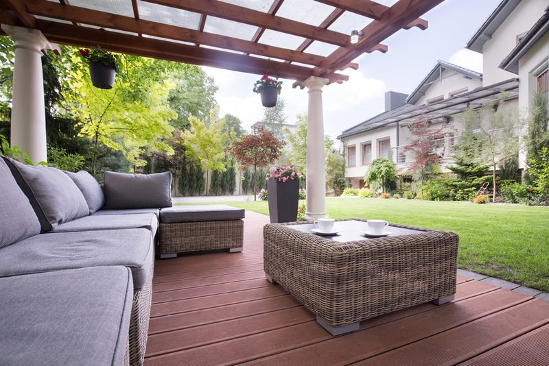 Outdoor patio with gray cushioned wicker seating and a matching coffee table under a wooden pergola, with lush lawn and a modern house in the background.