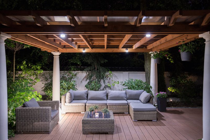 Night-time outdoor patio with a wicker sectional sofa and chairs under a wooden pergola, a glass coffee table, and surrounding potted plants.