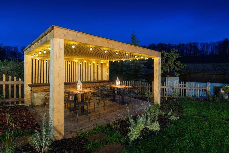 Wooden outdoor patio under a lit pergola with string lights, metal chairs around long tables, lanterns on the tables, garden and fence at dusk.