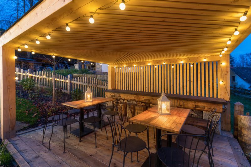 Outdoor wooden patio under a pergola with string lights, two wooden tables, metal chairs, lantern centerpieces; warm glow against a blue dusk sky.
