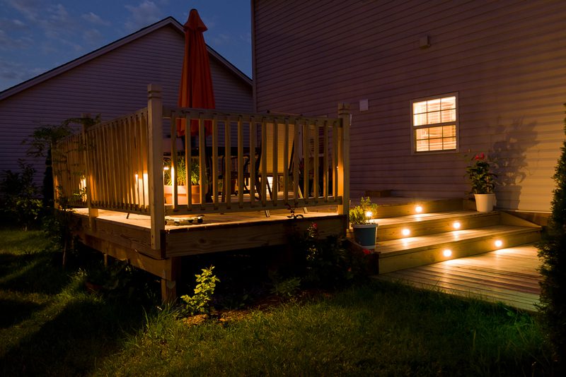 A wooden backyard deck at night with warm step lights, railing, a closed umbrella, outdoor furniture, potted plants, and a lit house window.