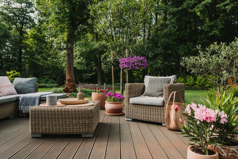Outdoor wicker seating on a wooden deck with cushioned sofas, a coffee table, potted pink and purple flowers, and a lush green garden backdrop.