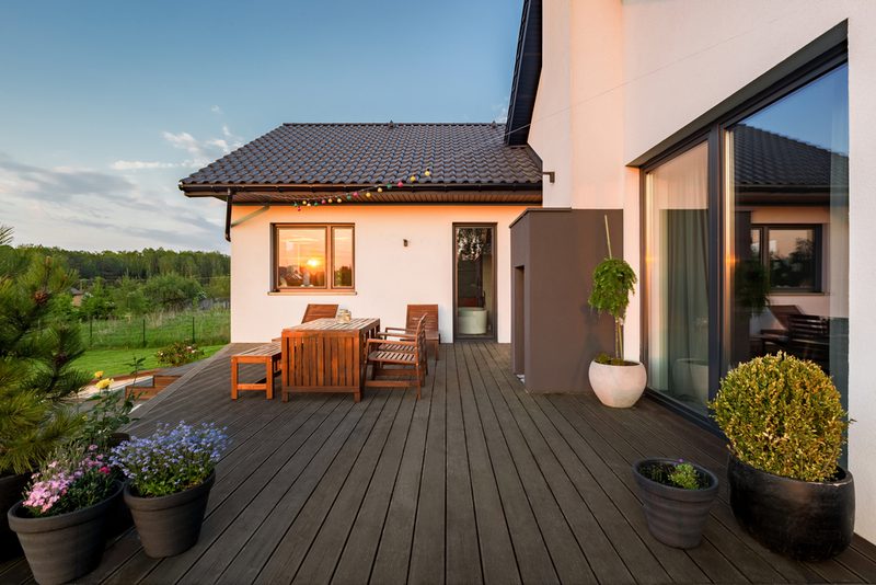 Modern white house with a large wooden deck, outdoor dining set, string lights, and potted plants; sunset glow in the window and a green lawn beyond.