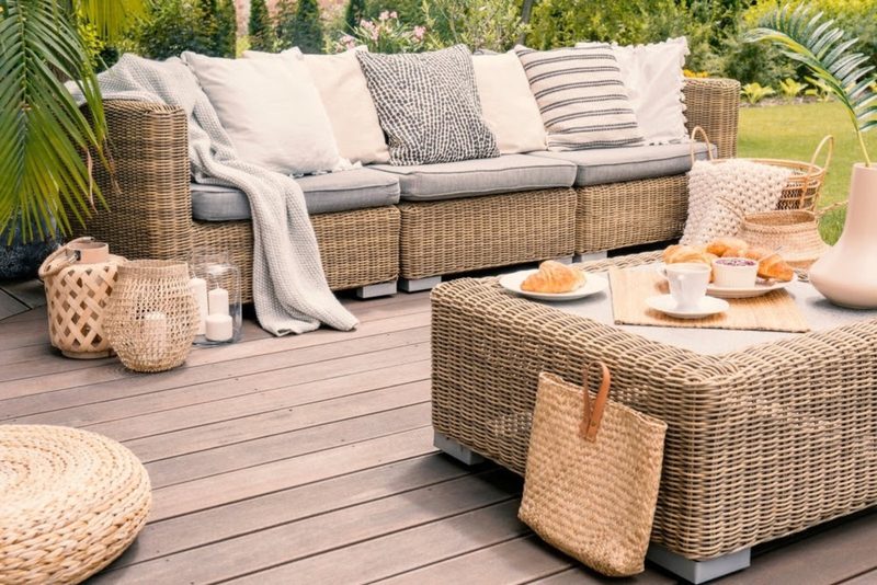 Cozy outdoor wicker seating area with a cushioned sofa, striped and neutral pillows, a woven coffee table, croissants and tea, baskets, and greenery.